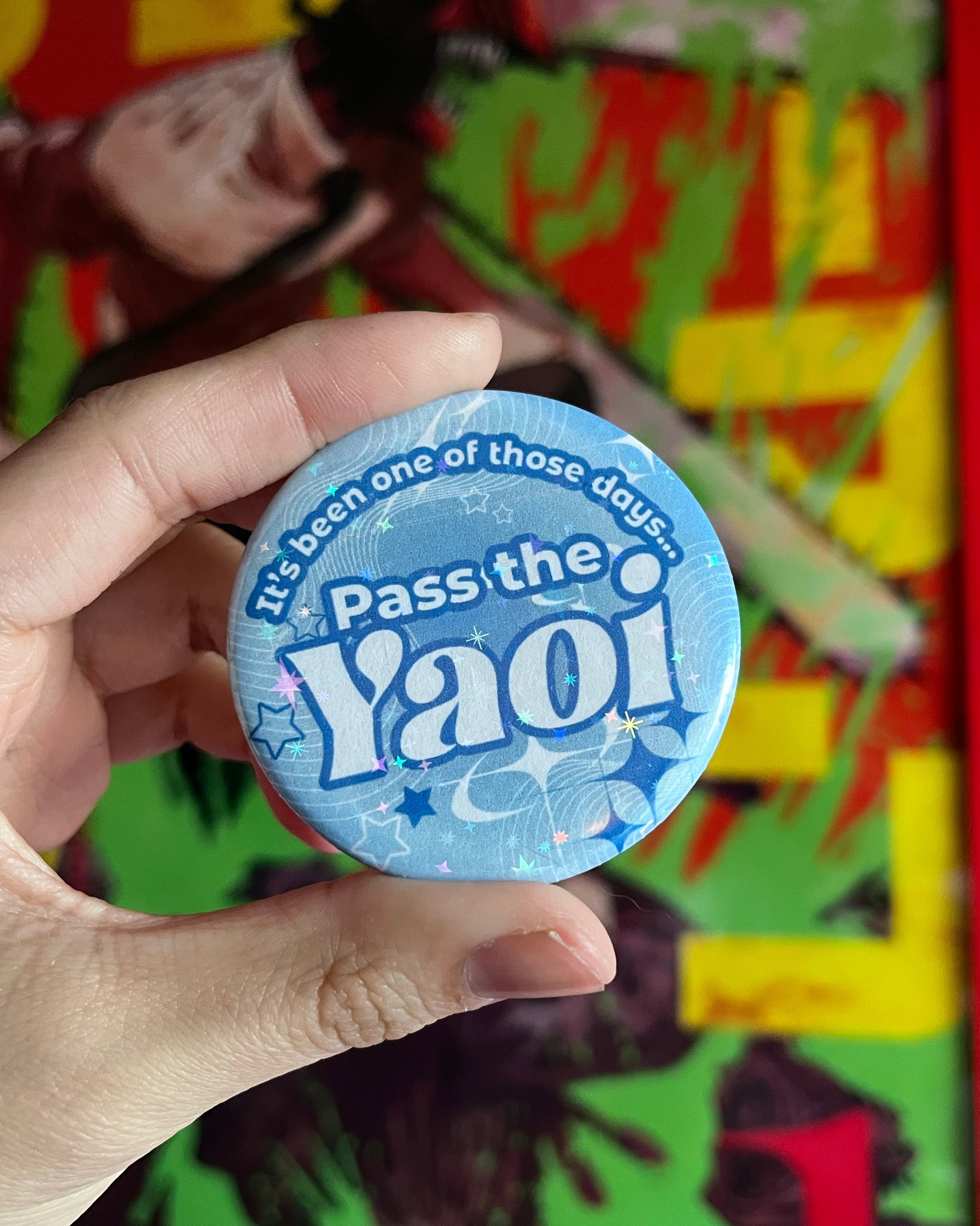 Hand holding a 'Pass the Yaoi' button in front of a colorful comic book background
